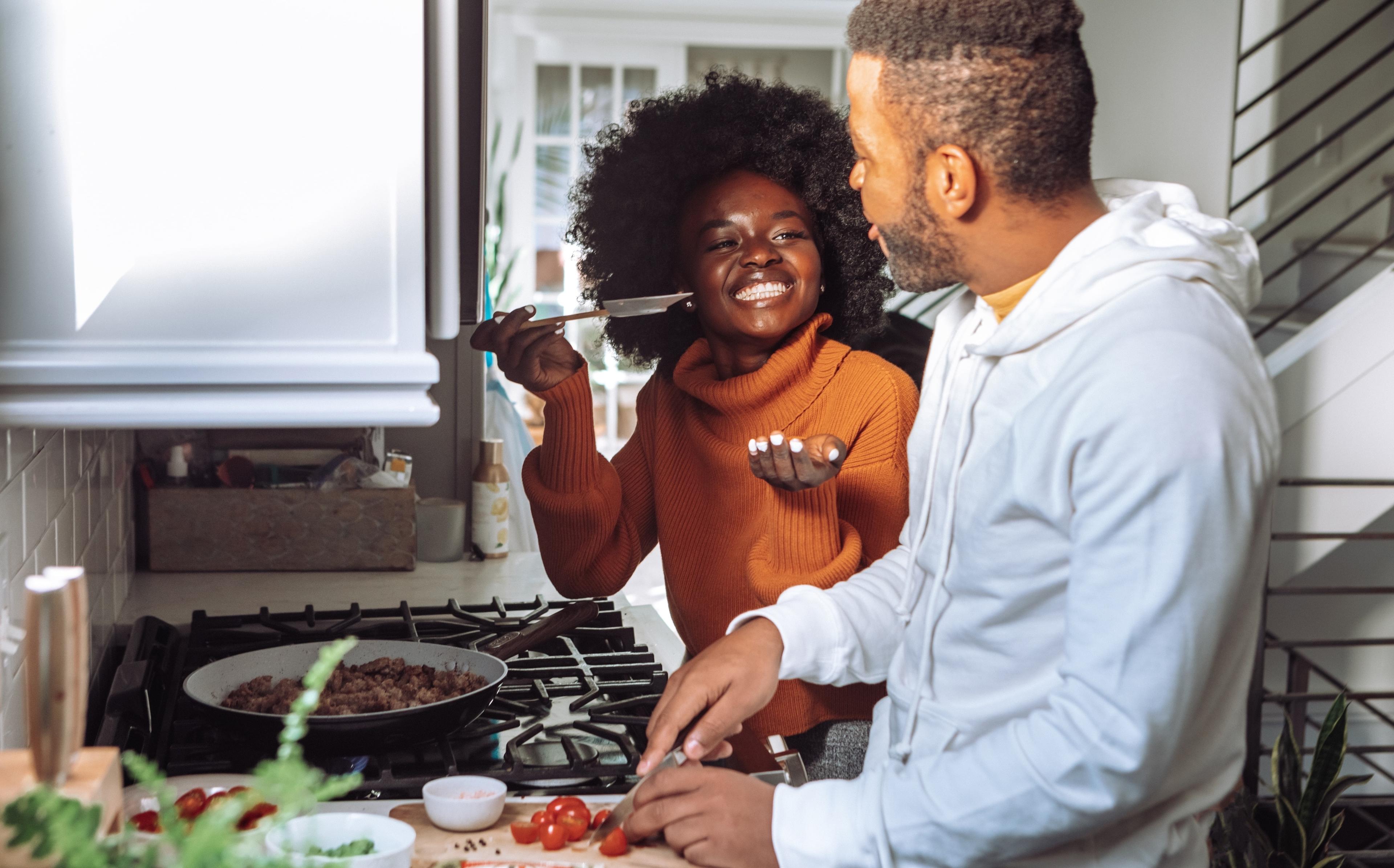 A man and a woman stand smiling in front of their stove while cooking ground meat and chopping tomatoes.