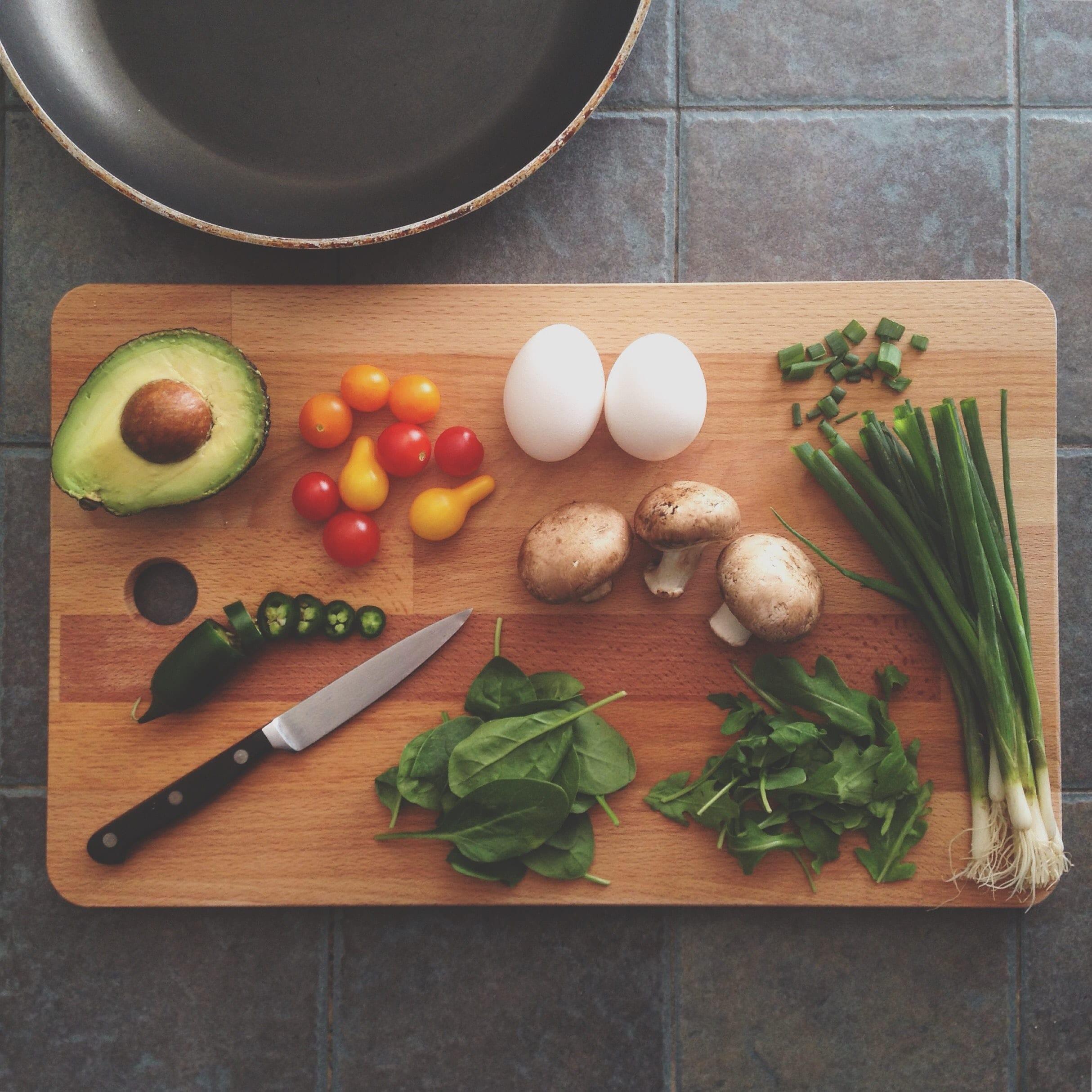 A cutting board witha knife, jalapeno pepper, avocado, cherry tomatoes, eggs, mushroom, chives and spinach.