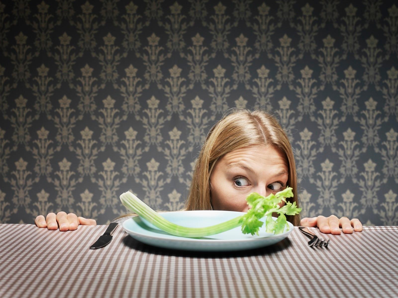 A woman stares skeptically at a piece of celery on an otherwise empty plate.