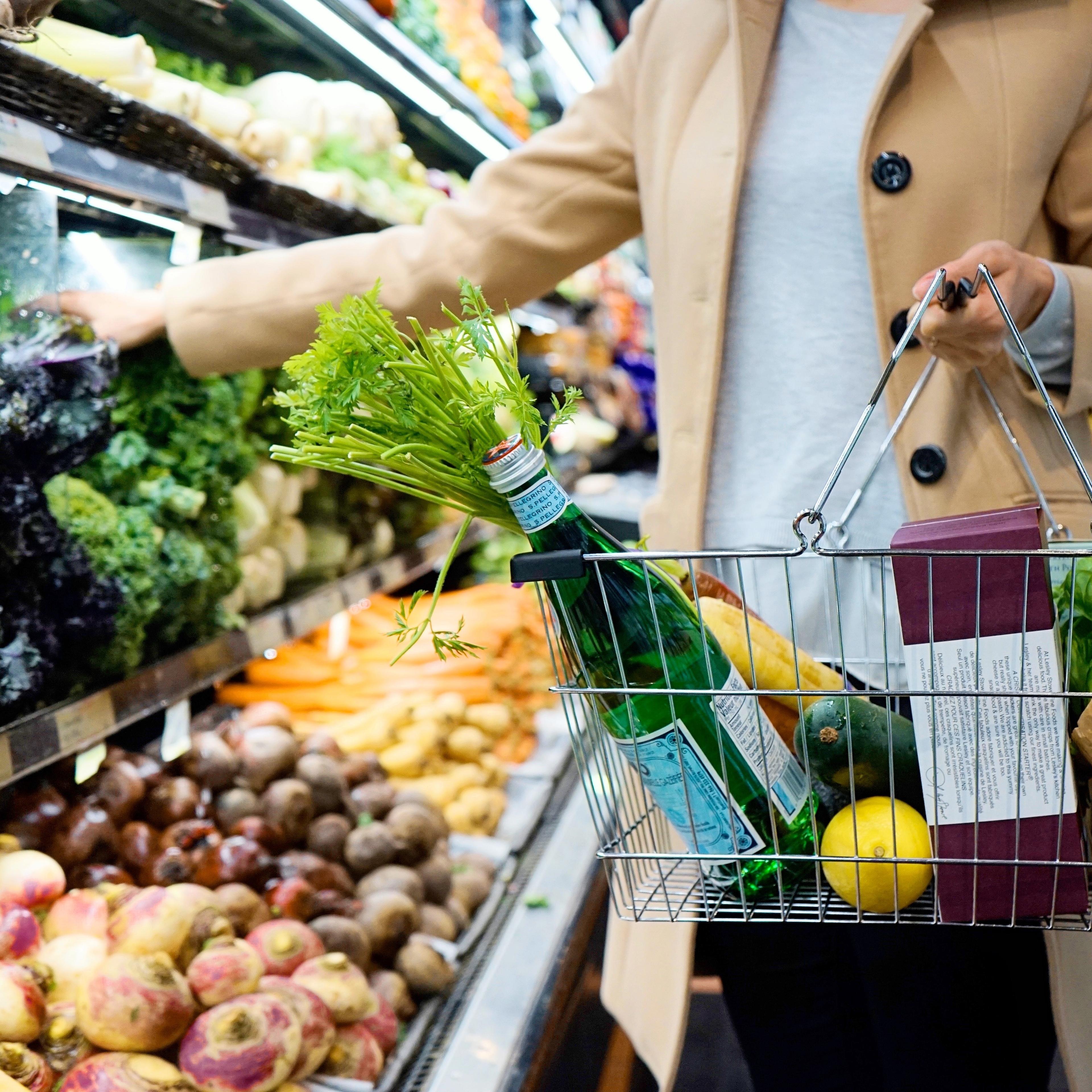 A person in a grocery store carrying a shopping basket with vegetables reaching for kale.