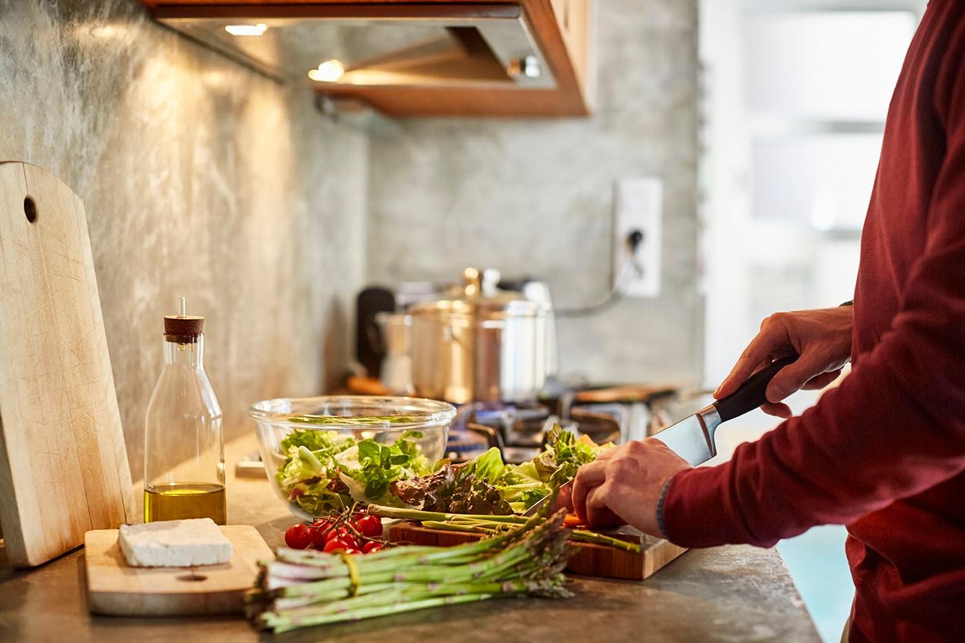 A person in a red shirt chops vegetables on a cutting board. On the counter is lettuce, tomatoes, zucchini, olive oil and a block of feta cheese.