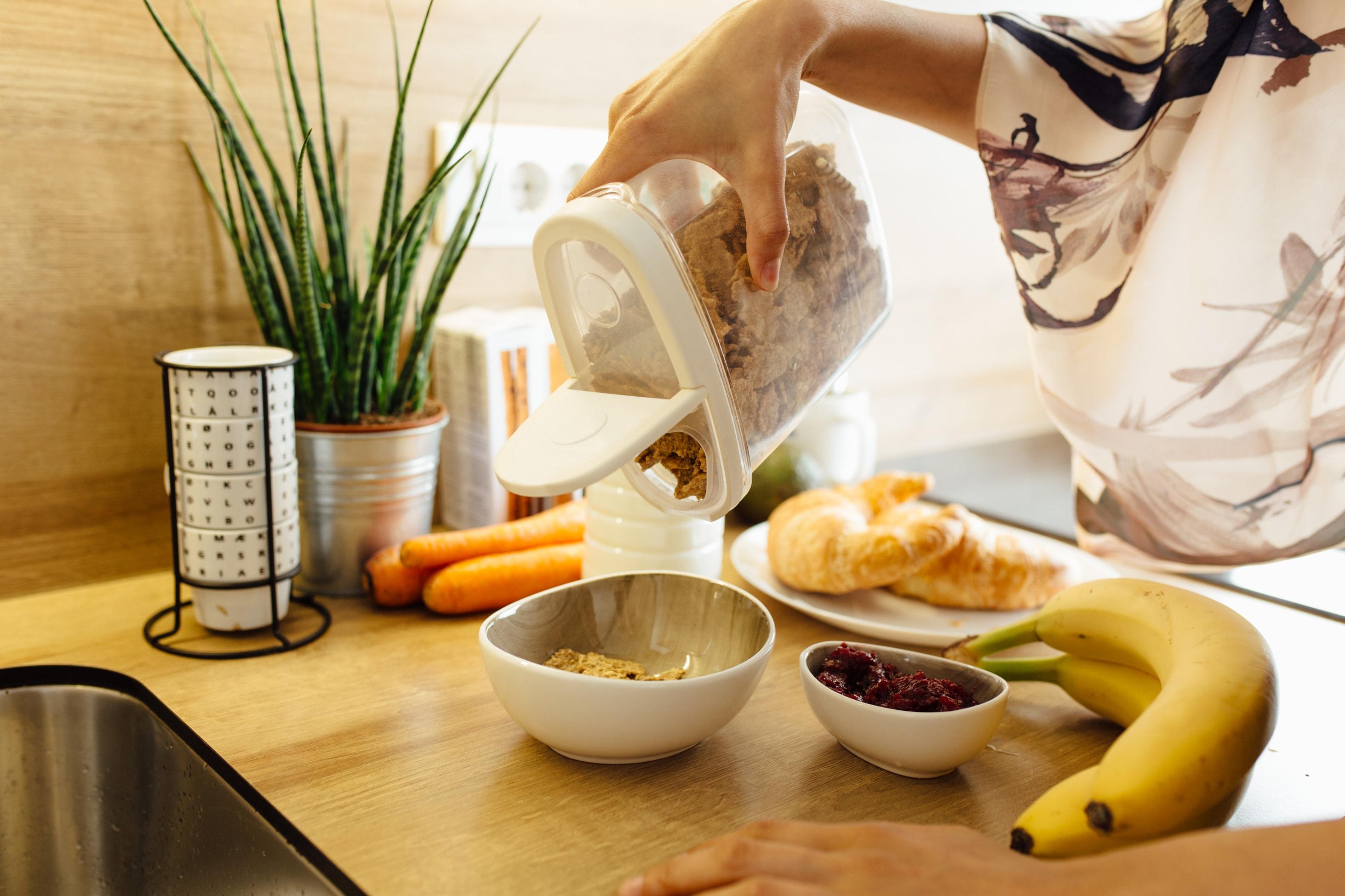 A woman stands in her kitchen pouring granola into a cereal bowl next to dried cranberries, bananas, carrots and croissants.