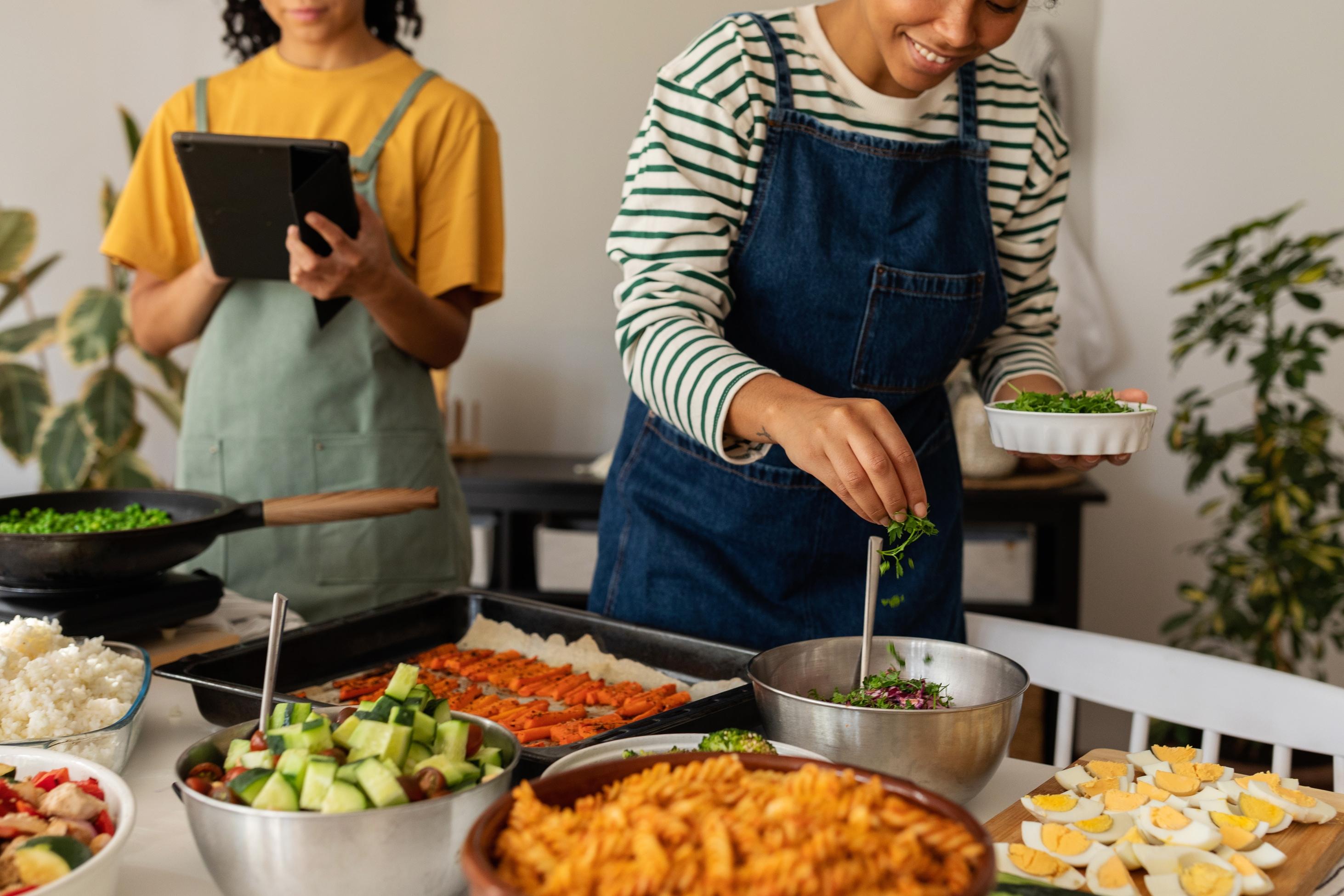 Two women, one holding a tablet, the other holding parsley, smile while cooking at a kitchen counter filled with salad, roasted red peppers, sautéed vegetables and eggs.