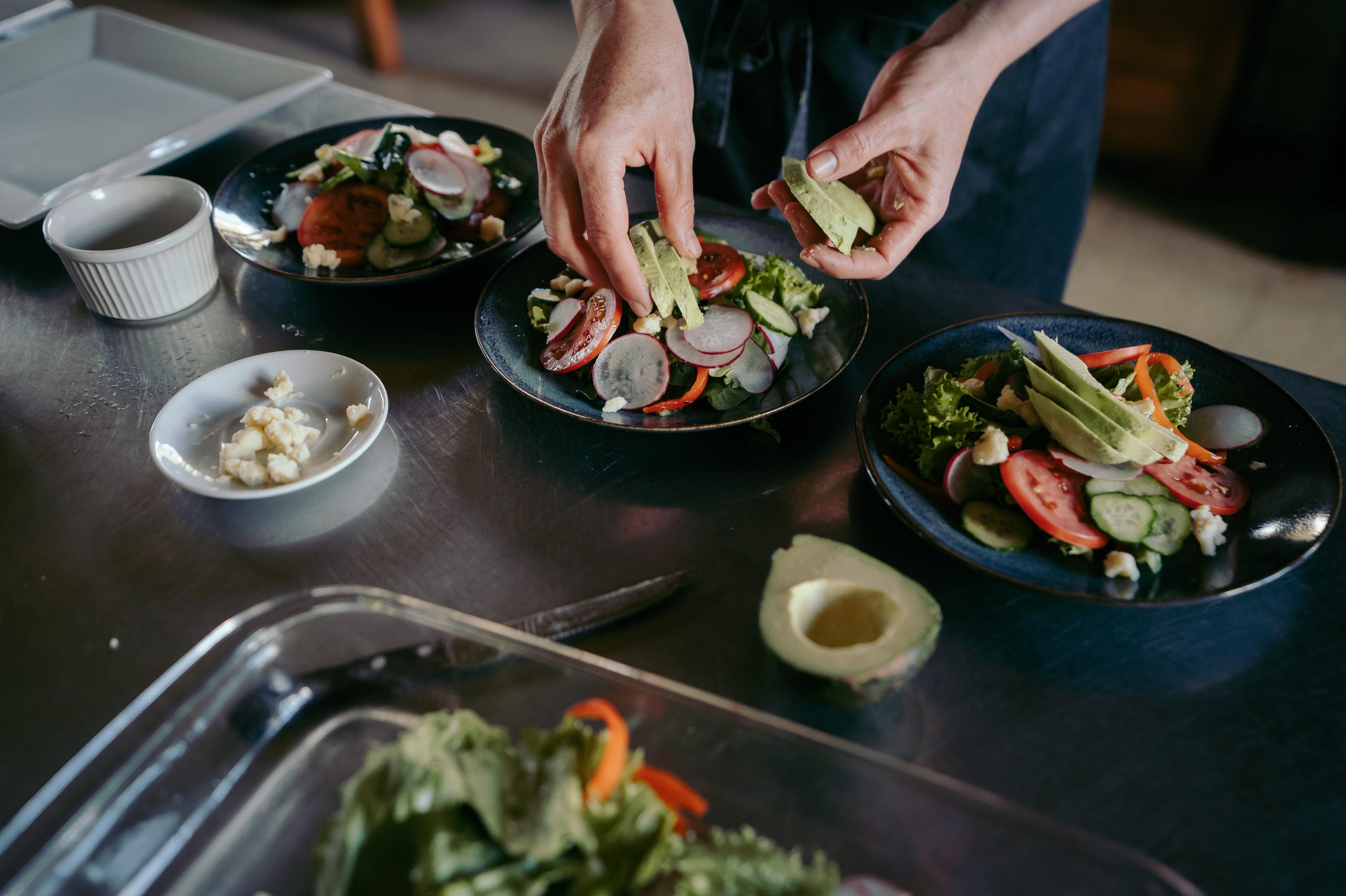 Une personne place des tranches d’avocat dans trois bols à salade bleus remplis de légumes verts, de tomates, de poivrons rouges, de navet et de fromage.